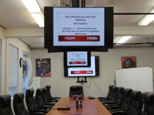 A classroom containing a long table, chairs, a dry-erase board, and three overhead screens displaying the name of an Aircraft Rescue and Firefighting course.