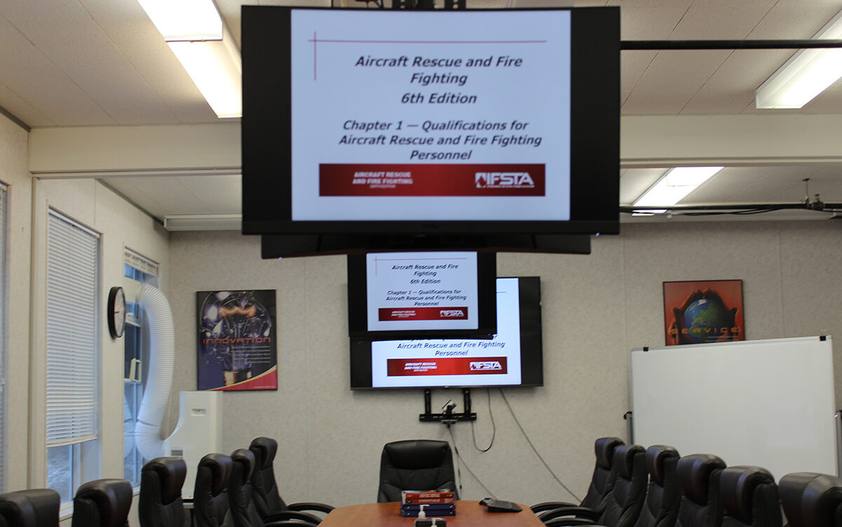 A classroom containing a long table, chairs, a dry-erase board, and three overhead screens displaying the name of an Aircraft Rescue and Firefighting course.